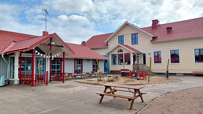 Skolgård med bänk i förgrunden, två skolbyggnader med röda tak i bakgrunden.