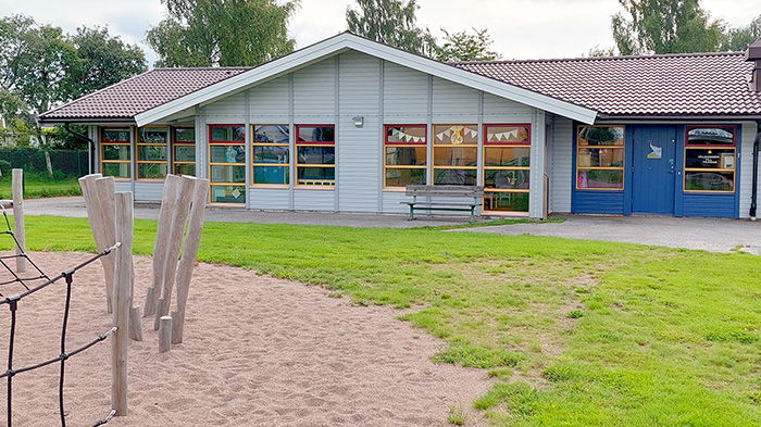 Del av skolgård i förgrunden, enplanshus med stora fönster i bakgrunden.